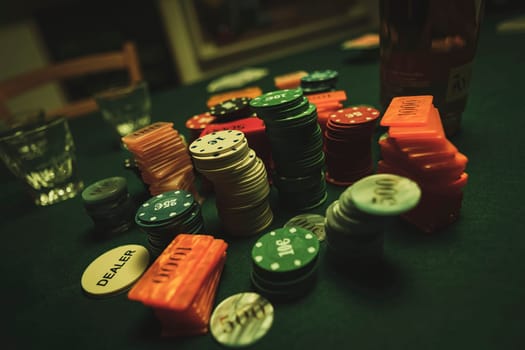Colorful poker chips and dealer button lie on green felt casino table with a bottle of liquor and shot glasses suggesting gambling night