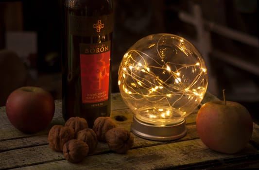 Luminous sphere casts a warm glow on a bottle of cabernet sauvignon, apples, and walnuts arranged on a rustic wooden table, creating a cozy and inviting atmosphere