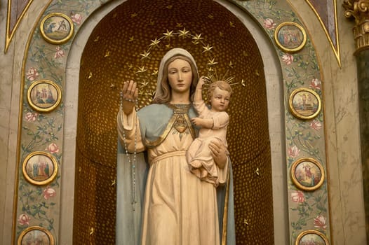 Statue of virgin mary holding baby jesus and a rosary, in front of golden background with stars and religious medallions