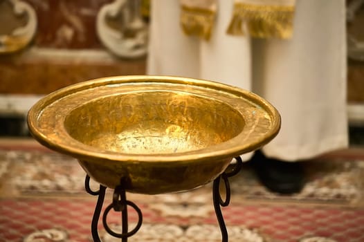 Golden baptismal font standing on a metal stand in a church, ready for a baptism ceremony with a priest in the background