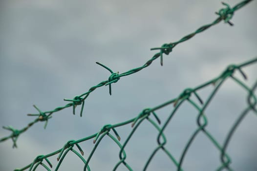 Close-up of green barbed wire atop a chain link fence, suggesting security and restricted access against a cloudy sky
