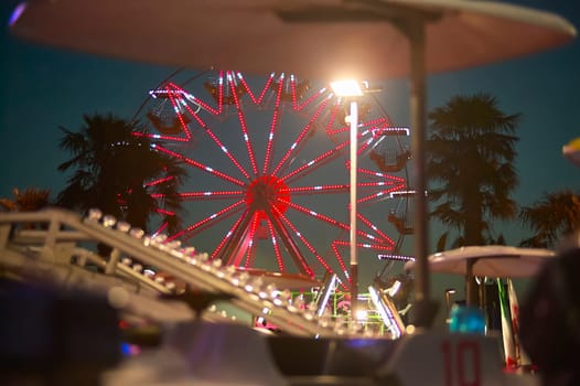 Red illuminated ferris wheel rotating at night in amusement park with palm trees and other attractions