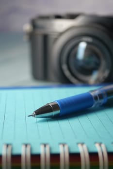 A stylish pen elegantly rests on an open notebook, with a camera artfully blurred in the background