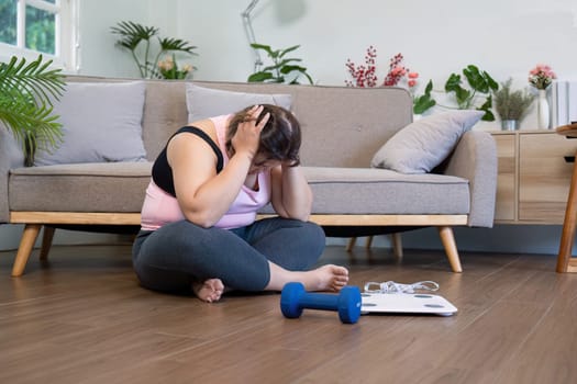 A woman sits on the floor feeling stressed about her weight loss journey surrounded by fitness equipment and a scale.