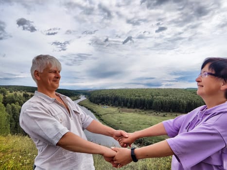 A couple shares a happy moment, holding hands and smiling as they overlook a peaceful river valley under a cloudy sky.