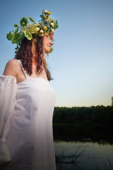 Slavic plump chubby girl in long white dress on the feast of Ivan Kupala with flowers and water in a river or lake on summer evening