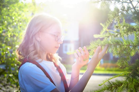 Young girl with glasses examines green tree branches, enjoying a sunny day in a residential area filled with greenery.