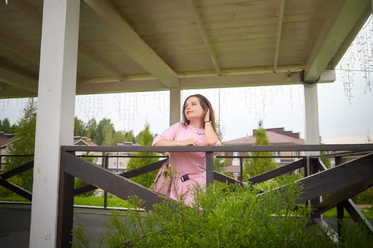 A young woman with long brown hair is wearing a pink shirt and is leaning against a white post