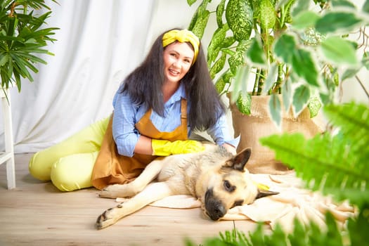 A gardener smiles while caring for flowers alongside a relaxed dog in a greenhouse.