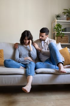 Front view of man comforting depressed wife sitting on the couch at home living room. Vertical image. Lifestyle concept.