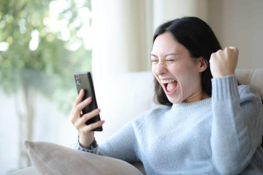 Excited asian woman celebrating success checking phone sitting on a couch at home
