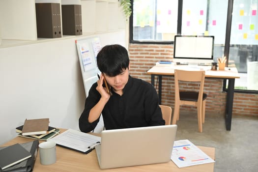 Young businessman having pleasant phone conversation and using laptop at his office desk.