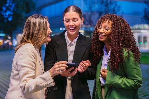 Three professional businesswomen of different ages and styles smiling while looking at a smartphone together in an urban nighttime setting.