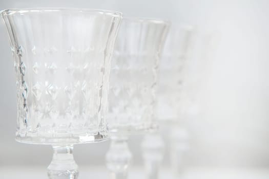 Crystal wine glasses with expanded edges and faceted pattern stand in a row on a white background, the first one is in focus on the sides, the second ones stand behind it in focus, close-up, the legs of the glasses are not visible. High quality photo