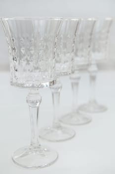 Crystal wine glasses on a long stem, with expanded edges and a faceted pattern stand in a row on a white background, the first one is in focus on the sides, the second ones stand behind it in focus. High quality photo
