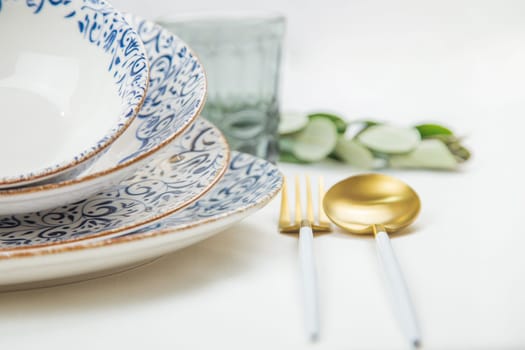 On the side there is a set of plates of different sizes with a white middle and blue patterns on the side of the plate and a gold border, they stand on top of each other with a tilt, there are gold - white devices on the side, there is a glass and green branches of plants, a side view of a close-up on a white background. The concept of serving. The concept of eating. High quality photo