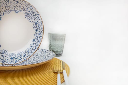 A white soup plate with a blue pattern on the edges stands directly on a blue-and-white patterned serving plate in close-up on a yellow straw napkin, next to cutlery are a knife and fork, on a white background. The concept of eating, table setting. High quality photo