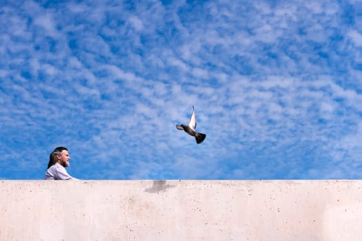 side view of positive mature male with beard resting near concrete fence and observing flying pigeon against cloudy blue sky, concept of freedom and middle age man lifestyle, copyspace for text