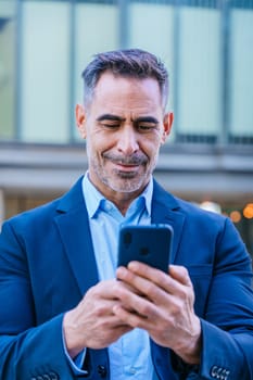 Smiling middle-aged businessman in a blue suit holding and using a smartphone outdoors in a modern urban setting.