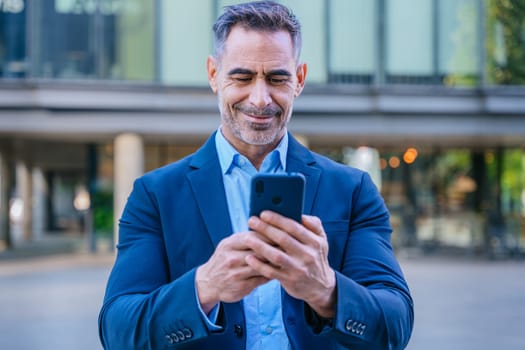 Smiling middle-aged businessman in a blue suit holding and using a smartphone outdoors in a modern urban setting.