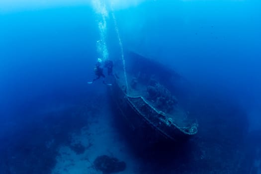 two scuba divers diving near of the wreck of an old ruined ship on the bottom of deep blue sea with clear water and light glare, concept of underwater adventure and seascape
