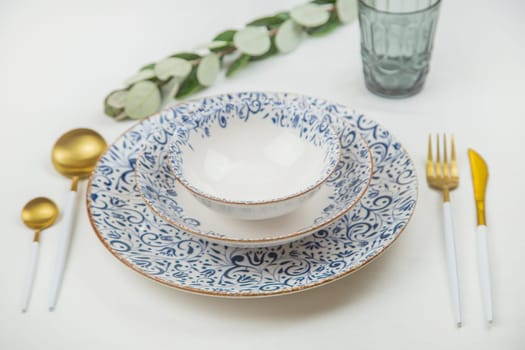 A set of white plates with a blue pattern on the edges stand on top of each other, spoons, a knife and a fork lie on the sides, behind them is a blue glass and a green sprig of sprouting on a white background. The concept of eating, table setting. High quality photo