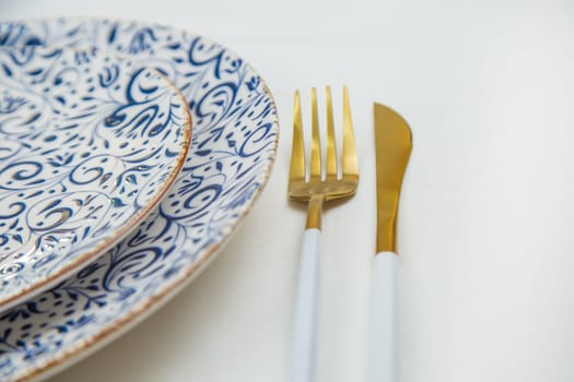 On the side, two serving plates with blue patterns on a white background stand one on one, part of the plates is visible, gold-white appliances are located on the side, a close-up side view on a white background. The concept of serving. The concept of eating. High quality photo