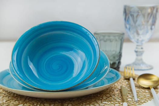 Bright blue serving plates with a spiral pattern, tilted one after the other, serving devices of gold - white color are located on the side, glasses and glasses are visible behind them, side view on a white background with a horizon line. The concept of serving. The concept of eating. High quality photo