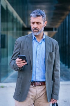 Smiling middle-aged businessman in a blue shirt holding and using a smartphone outdoors in a modern urban setting.