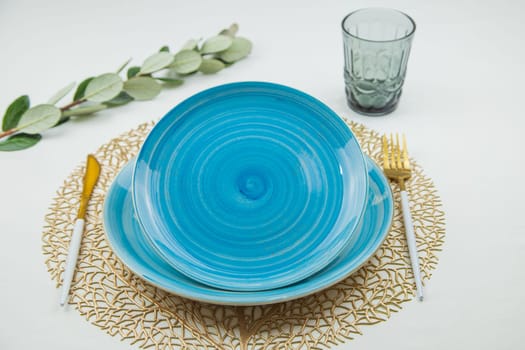 Bright blue serving plates with a spiral pattern, tilted one after the other, serving devices of gold - white color are located on the side, there is a glass on top and a green branch of the plant lies, top view on a white background. The concept of serving. The concept of eating. High quality photo