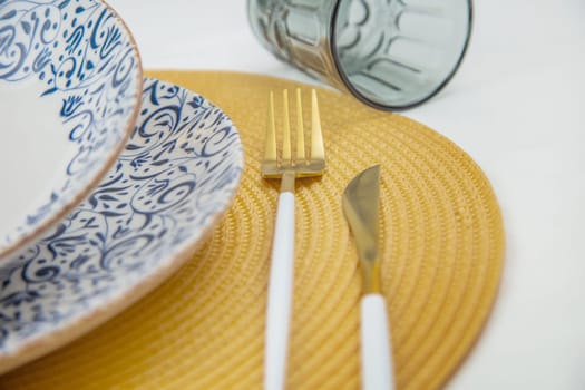 A white soup plate with a blue pattern on the edges stands on a blue-and-white patterned serving plate in close-up on a yellow straw napkin, next to it are cutlery, a knife and fork, behind them is a blue glass on a white background. The concept of eating, table setting.High quality photo
