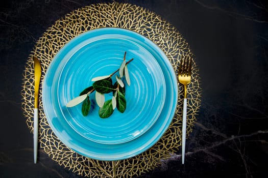 Green twigs lie on blue plates in the center, plates stand on gold patterned napkins, golden cutlery lies on the sides: a fork and a knife on a black background. The concept of serving, the culture of eating.High quality photo