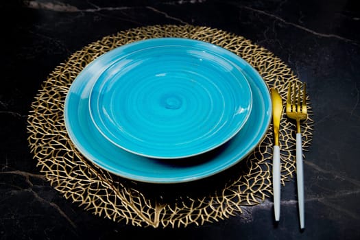 Blue plates stand on gold patterned napkins, gold cutlery on a black background, top view. The concept of serving, the culture of eating. High quality photo