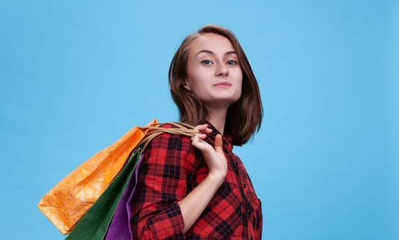 A cheerful young woman stands confidently against a blue backdrop, showcasing her vibrant shopping bags. She embodies the joy of the commerce season, ready for an exciting day.