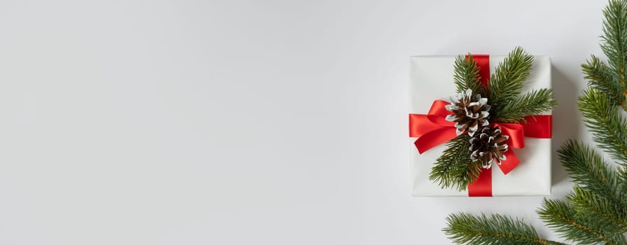 Banner with a white gift box with a red ribbon and fir twigs and cones on white background. Space for text.