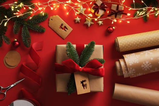 Festive gift wrapping scene with garland lights. Kraft brown paper rolls, red velvet ribbon, labels, scissors and finished wrapped present.