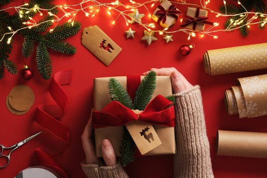 A woman's hands hold a wrapped gift. Table with handmade items, rolls of kraft brown paper, red velvet ribbon, labels, scissors.