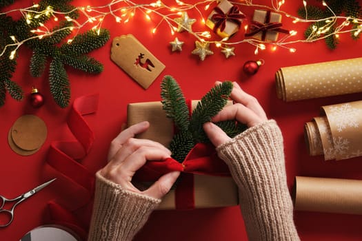 Women's hands wrapping a gift. A table with handmade items, rolls of kraft brown paper, red velvet ribbon, labels, scissors.