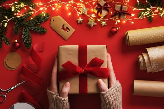 A woman's hands holding a gift. Table with handmade supplies, rolls of kraft brown paper, red velvet ribbon, labels, scissors.