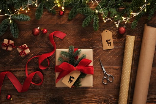 Festive gift wrapping scene with garland lights. Kraft brown paper rolls, red velvet ribbon, labels, scissors and finished wrapped present.
