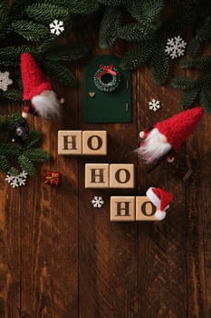 Wooden blocks with Ho-Ho-Ho text in English with Christmas tree branches, gnomes in red hats, door with wreath, Santa hat. Space for your text.