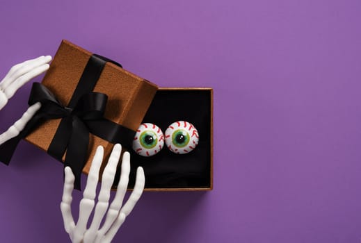 Halloween skeleton hands holding a gift box with fake eyeballs on purple backdrop. Background for Halloween cards, banners, posters. Place for your text.