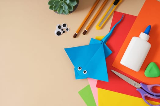 Work table for paper crafts colored sheets, glue, scissors, pencils, stationery knife. Finished origami toy stingray. Beige background, space for your text.