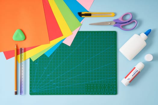 Origami kit on green cutting mat. Colored paper, scissors, knife, glue, pencil, eraser, ruler. Space for text.