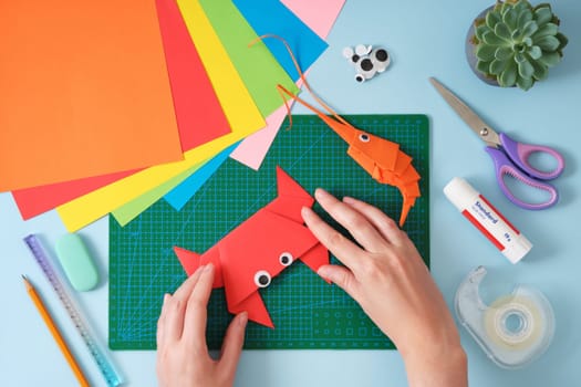 Top view of paper cutting tools, scissors, cutter and paper crafts. Women's hands fixing the final folds for the crab toy. DIY project concept for kids. Flat lay