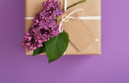 Brown craft gift box with empty tag, lilac flowers on violet background. Place for your text