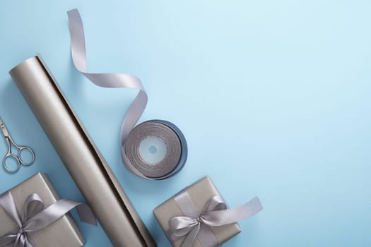 Wrapping gifts in gray craft paper with silver ribbon on light blue background top view, copyspace