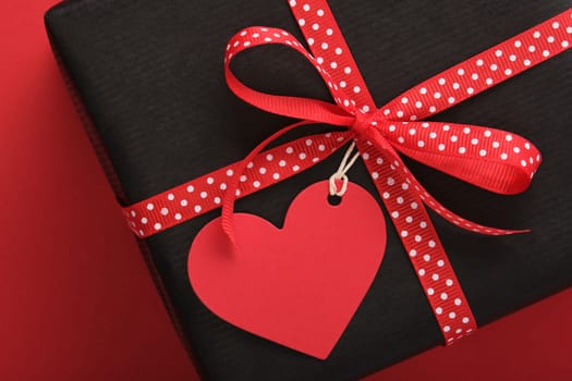 Card with black craft gift box with empty heart-shaped label on red background. Space for your text