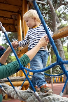 Little boy holding his mother's hands as he walks across the bridge on the playground. Vertical shot