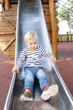 A little blonde boy slide down a slide on the playground. Vertical shot